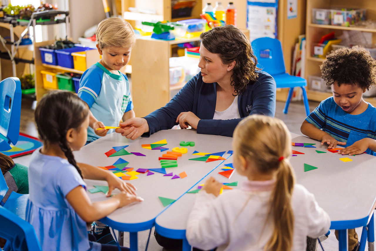 Teacher at a table of children creating with colorful shaped paper in a preschool classroom